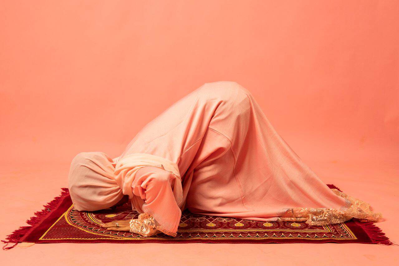 services-02 A woman in a pink abaya praying on a decorative prayer rug in a studio setting.