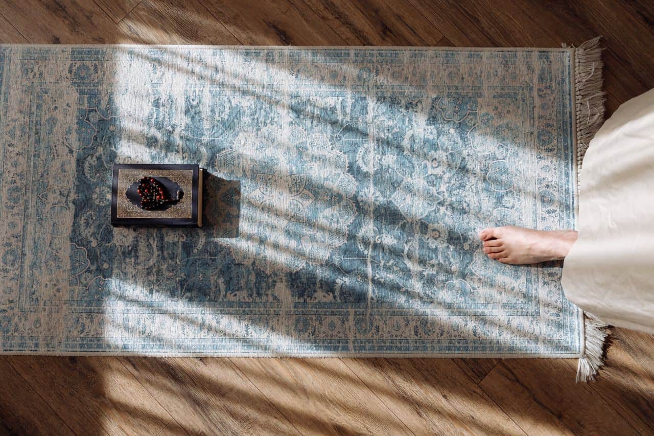 services-01 Top view of a prayer setup with Koran and beads on a patterned rug, sunlight casting shadows.