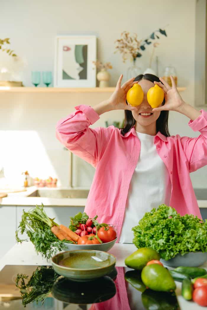A cheerful woman playfully holds lemons over her eyes in a bright kitchen with fresh vegetables.